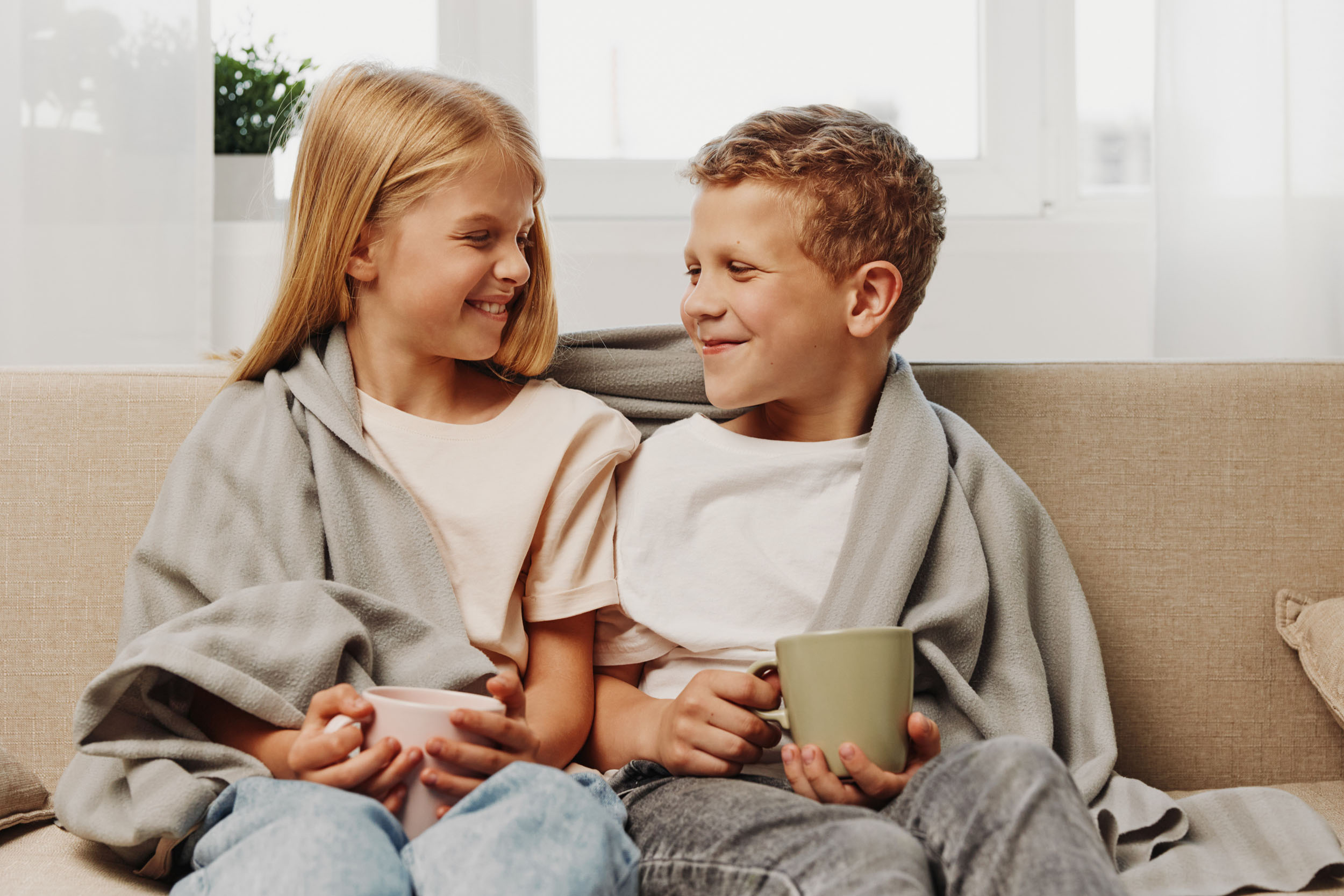 Wärmedämmung – Peter Weigert Mädchen und Junge sitzen lachend in eine Decke gekuschelt auf dem Sofa und haben Tassen in der Hand